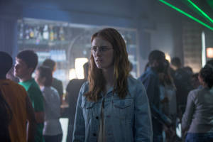 戴眼鏡、身穿牛仔外套的年輕女子夜晚站在擁擠的酒吧裡。 — Netflix GIF 壁紙