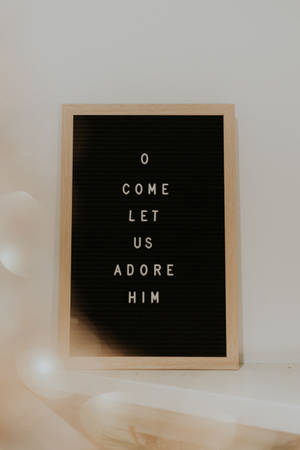 一個木框字母板，白色文字寫著「O COME LET US ADORE HIM」，放在淺色牆上。